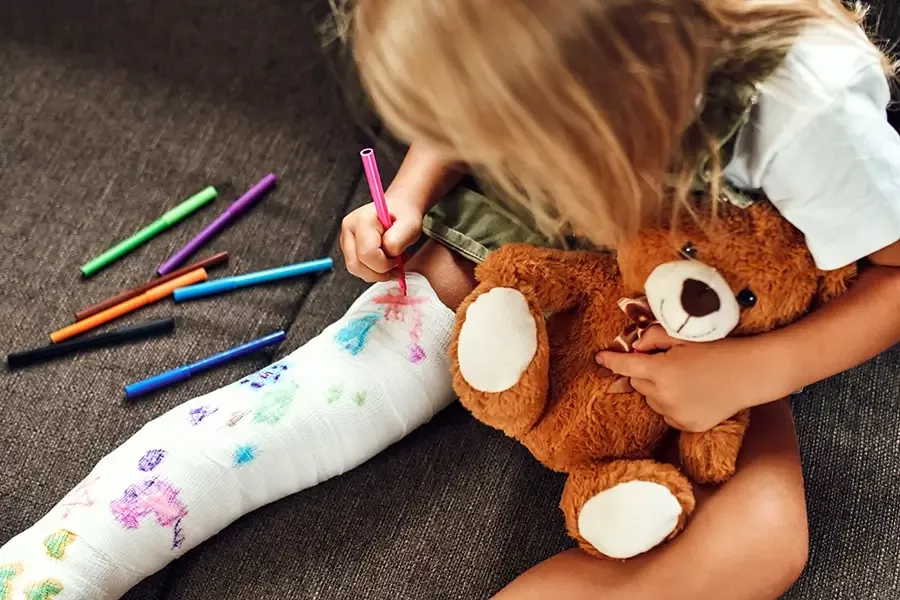 child coloring a cast on her leg holding a teddy bear