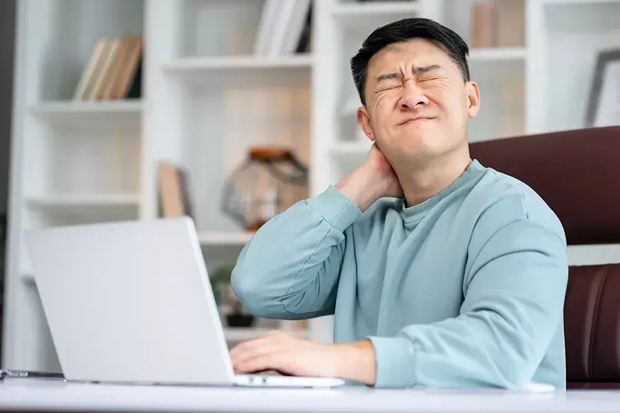 Photo of a man holding his neck and grimacing in pain while working on a laptop